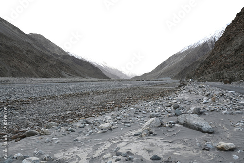 A wide, remote mountain valley landscape in Ladakh, India, with a dry, rocky riverbed and distant snow-capped peaks.