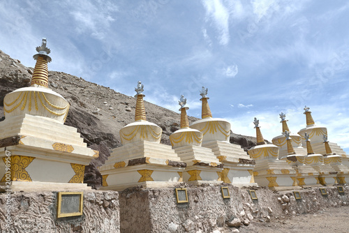 A row of traditional white and gold Buddhist stupas against a rocky mountain backdrop.