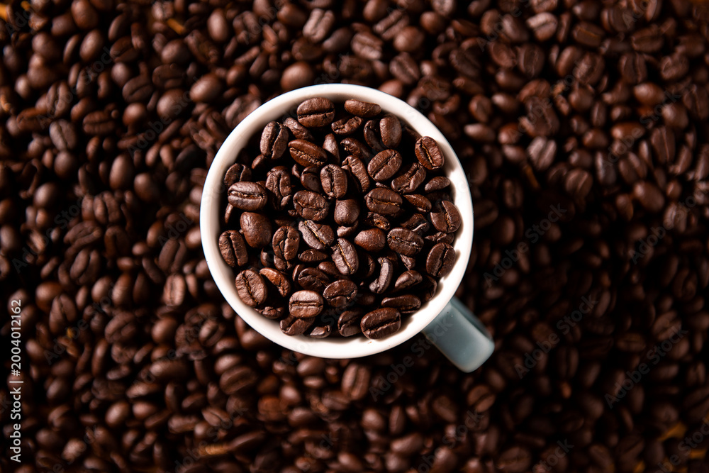 Fototapeta premium Top down view of a white ceramic cup filled with aromatic dark roasted coffee beans on a background of scattered beans for cafe and breakfast concept