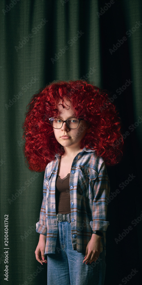 Obraz premium Girl with oversized red curly hair and glasses stands against dark green curtain. Concept of bold individuality, self-expression, growing confidence and feeling larger than life.