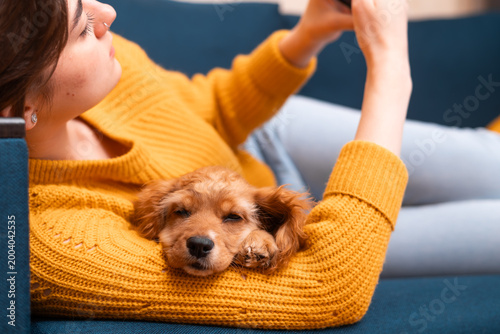 A woman lies on a couch, engaged with her smartphone while a small dog rests on her arm. The scene reflects a casual moment of companionship between the woman and her pet