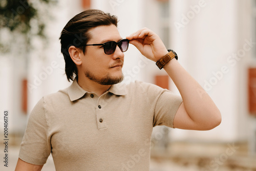 Fotografi Pensive Man Lifting Sunglasses Beside Architectural Facade, Polo Shirt, Tiedback