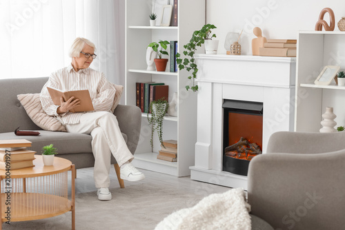 Senior woman in eyeglasses reading book on sofa near fireplace at home