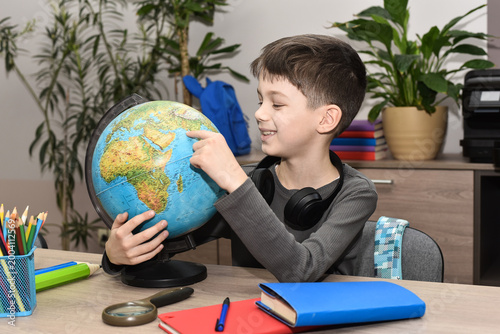 Happy school-age boy sitting at a table with a globe, exploring geography and studying at home.