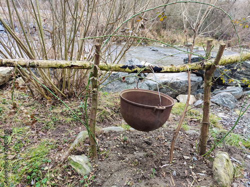Картината върху платно Old empty tourist cauldron suspended above campfire area on riverbank