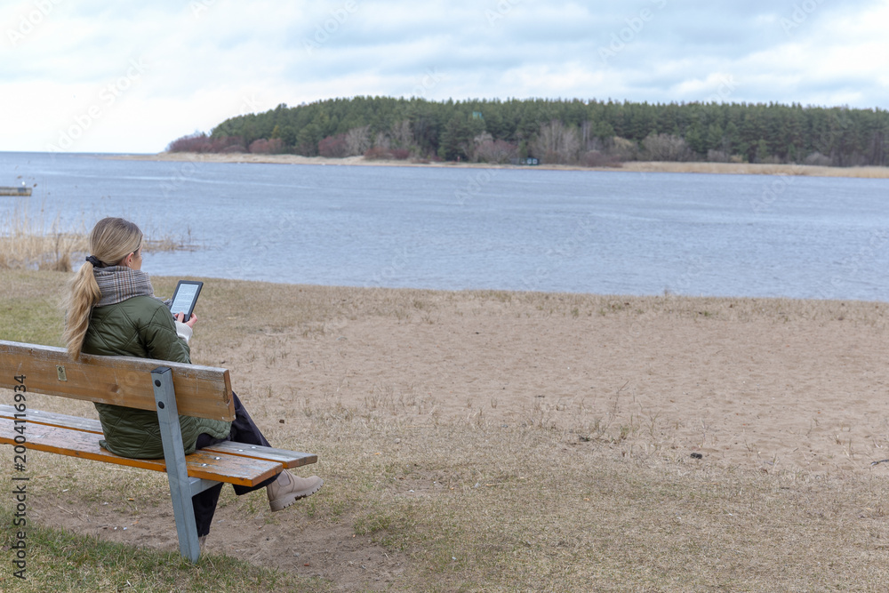Fototapeta premium Woman sitting on bench by lake using tablet, remote work and reading outdoors, calm lifestyle and digital lifestyle concept