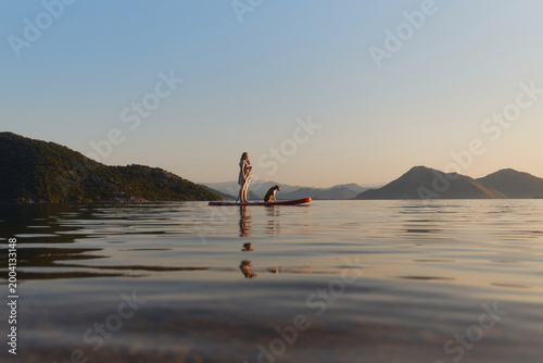 A woman stands with dog on a paddle board during dusk. The scene is quiet with smooth water and distant mountains.