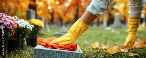 A person wearing yellow gloves cleans a gravestone with an orange cloth in a cemetery filled with autumn leaves and flowers.
