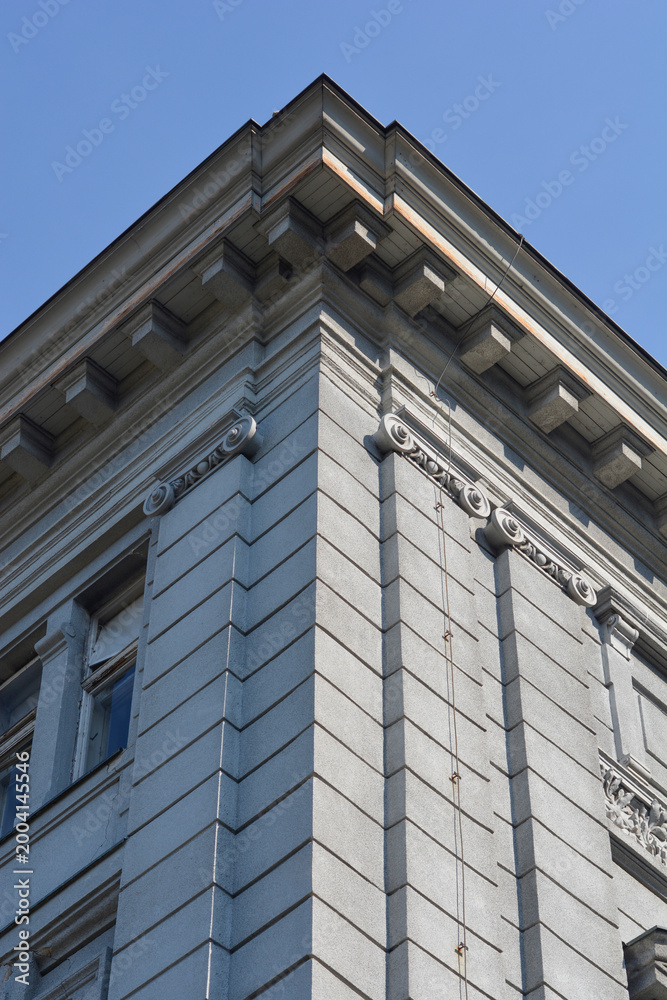 Obraz premium A low angle view of a grey classical building corner featuring decorative cornices and ionic order details against a clear blue sky