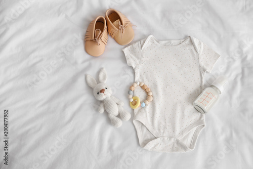 Bottle of baby milk formula, toys, booties and bodysuit on light bed