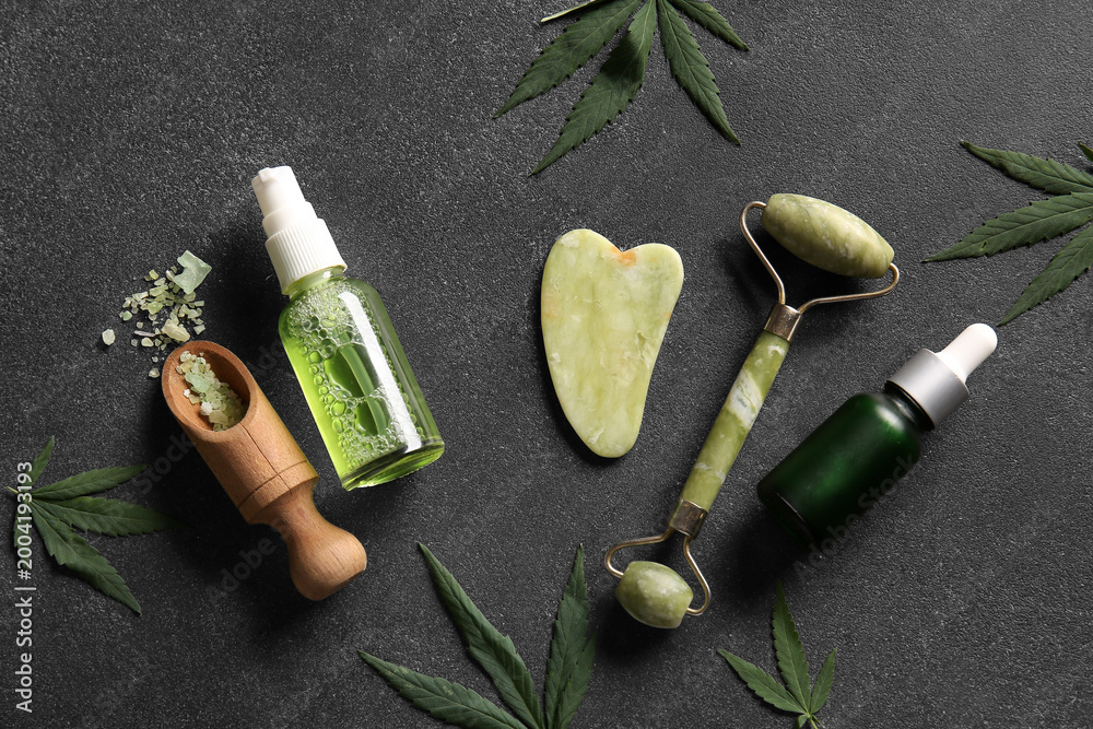 Obraz premium Bottles of hemp cosmetic products with sea salt, facial massage tools and leaves on black background