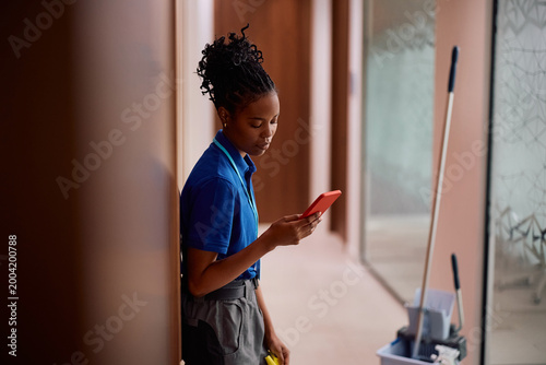 Fotografie Black woman using cell phone while working as professional cleaner in office building