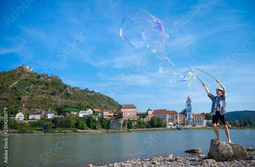 Happy young man making giant soap bubbles on the riverbank with the picturesque town of Dürnstein and castle ruins in the background, Wachau Valley, Austria.