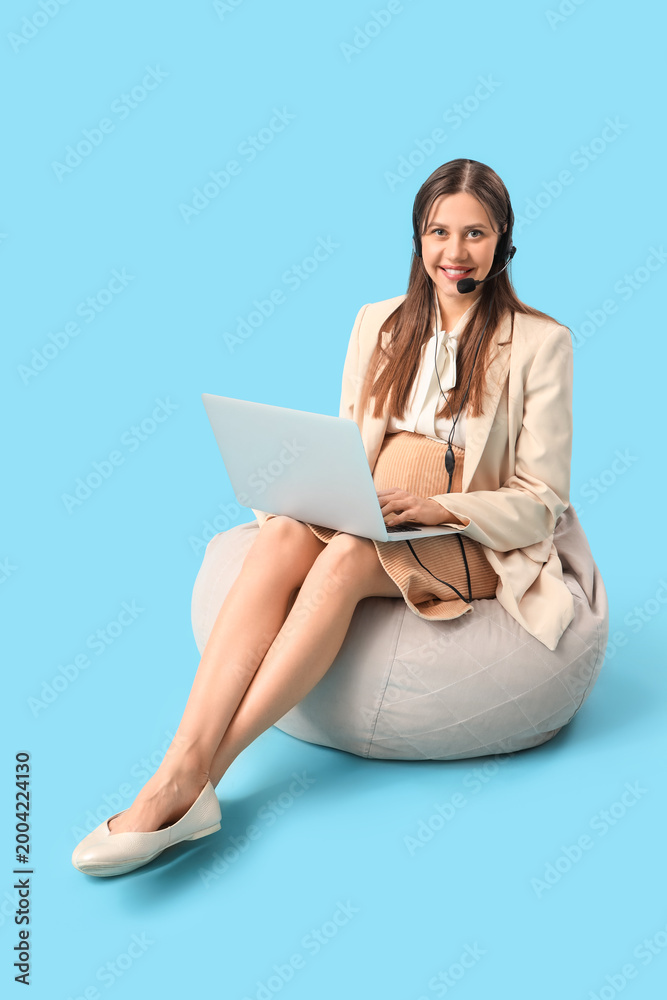 Fototapeta premium Young pregnant businesswoman in headset working with laptop on blue background