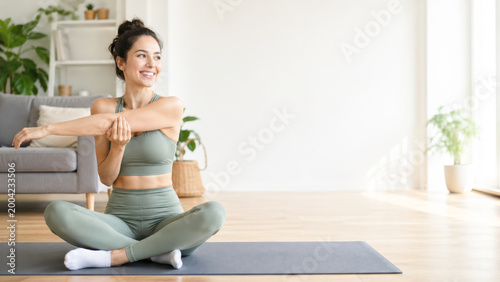 A woman in activewear smiles while stretching on a yoga mat in a bright, cozy living room.