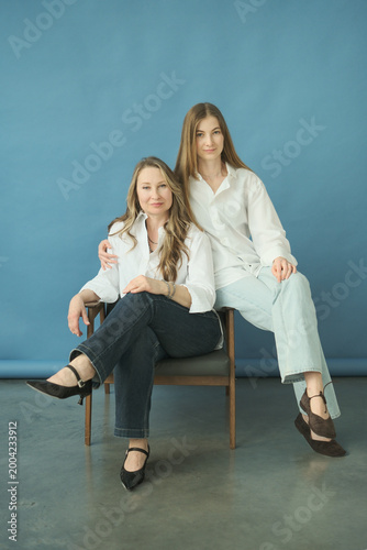 Beautiful mature mother and adult daughter in white shirts and jeans hugging and sitting on chair in the photostudio on blue paper background. Two female generations. Mother's day concept