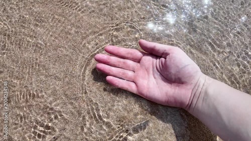 Close-up of a hand touching clear sea water and sand on a sunny beach. Concept of relaxation, tropical vacation, mindfulness, and connecting with nature.