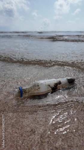 Dirty plastic bottle washed up on the sandy beach by waves. Concept of ocean pollution, and environmental conservation. 4K vertical slow-motion footage.