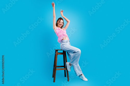 Bild auf Leinwand Happy young woman dancing on stool in pink top and jeans against bright blue stu