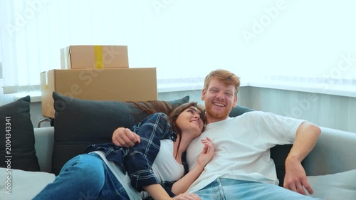 Camera approaching joyful male and pretty female smiling jumping on a sofa in light room with many cardboard boxes. Close up of happy boyfriend and girlfriend moving in together. Real time