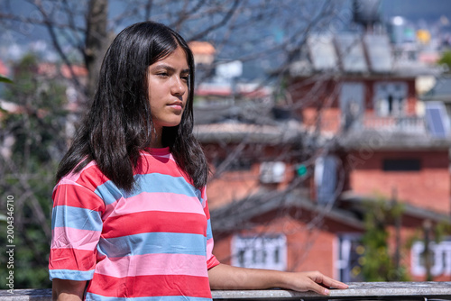 Asian woman in pink and blue striped top looking sideways on rooftop courtyard thoughtful expression