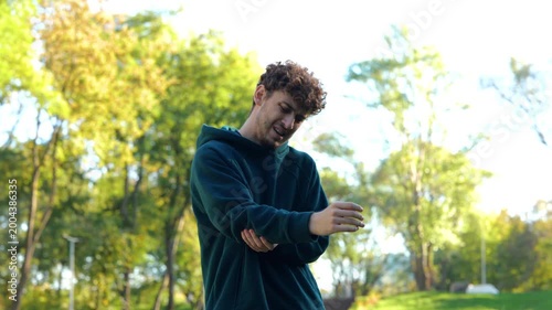 Sportsman running in park, feeling arm hurt after workout. Young Caucasian man standing outside on summer sunny day. Guy massaging his arm. Real time.