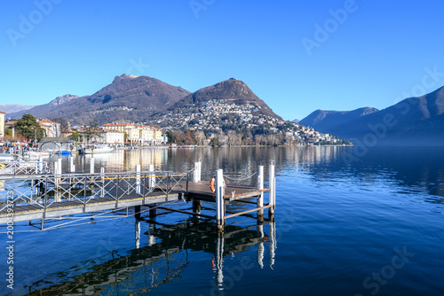 Peaceful waterfront scene in Lugano with a wooden pier, calm lake reflections, and mountain views under a clear blue sky, capturing serenity and Swiss travel charm.