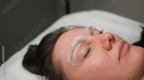 Professional cosmetologist applying a white cream for an eyebrow lamination procedure on a woman