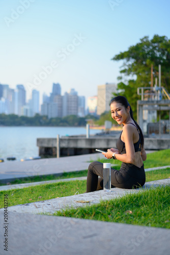 Sporty young woman smiles at camera holding phone after workout by city lake