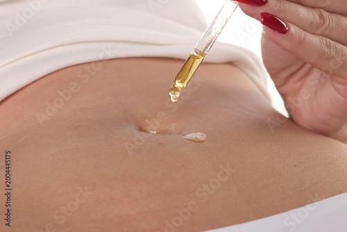Fotografie Close-up of a woman applying cosmetic oil with a dropper directly into her navel for skincare, studio shot on a white background