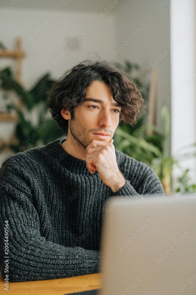 Fototapeta premium Young man pondering while using laptop in bright indoor room with plants