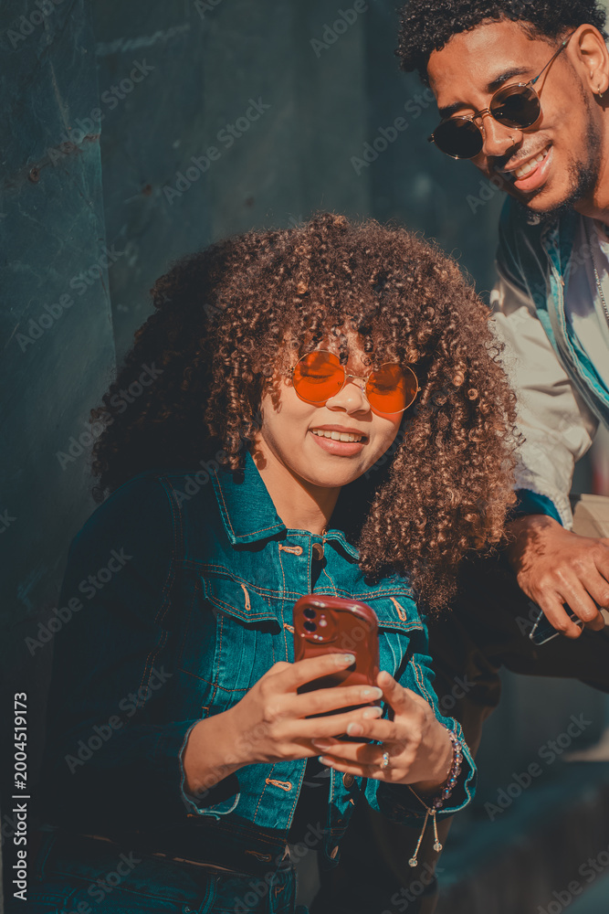 Fototapeta premium Young stylish couple sharing a fun moment looking at a smartphone outdoors, smiling in natural sunlight against a teal wall