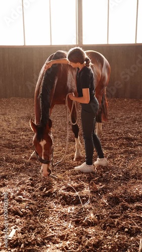 Teenage girl grooming, brushing carefully brushing brown horse's mane bay horse in indoor arena at sunset. Equestrian lifestyle, responsibility, animal care, bonding concept, teenage rider, hobby