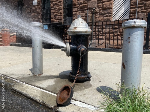 Black and silver fire hydrant with spray cap to conserve water spraying onto street on hot summer day in Harlem, New York City