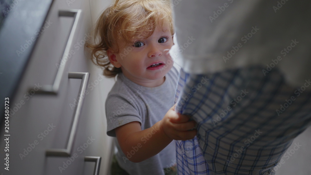 Fototapeta premium Toddler standing near mother in kitchen while she cooks, holding onto her clothes and seeking attention during candid authentic family moment at home