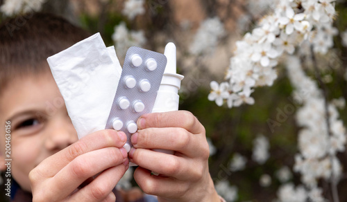 Child holding nasal spray, pills and tissue against a blooming tree. Allergy