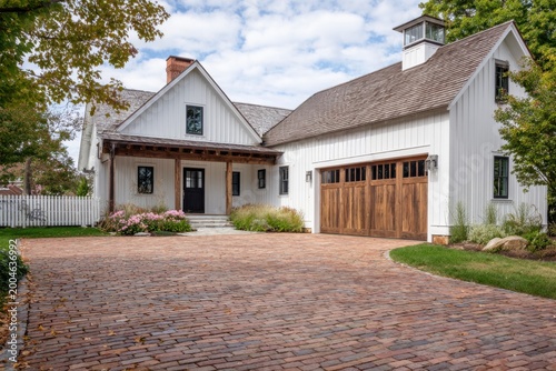 A contemporary white house features elegant wooden garage doors and is complemented by a brick driveway, all set beneath a bright blue sky adorned with fluffy clouds during the day