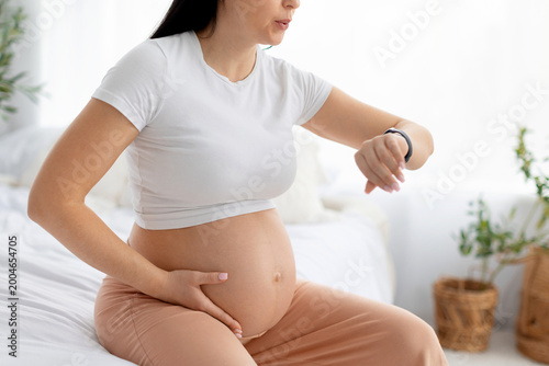 Pregnant woman sitting on bed checking smartwatch while holding belly, timing contractions or monitoring health data during late pregnancy at home in bright cozy bedroom