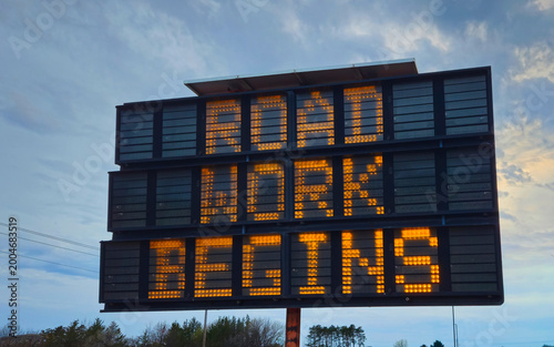 Electronic road sign displaying ROAD WORK BEGINS in bright orange text against a blue sky. Traffic control, infrastructure, construction, and safety themes. Design Element.