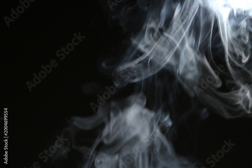 A white curved texture, magical smoke from an incense stick rising from the surface against a dark black background with copy space. Mystical pattern, concept of meditation and relaxation. Magical fog