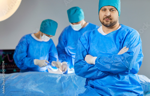 Surgeon confident man, group professional medical doctors surgical team performing surgery, doing operation, procedure on patient, wearing blue surgical gown, cap, face mask, treat injuries, diseases