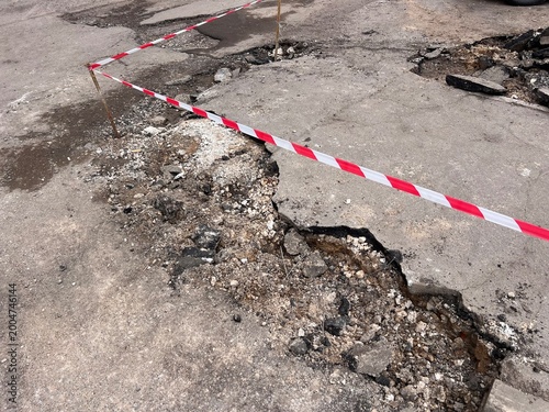 damaged asphalt road with large cracks and a hole, marked with red and white caution tape. Concept of poor infrastructure, road repair, construction work, and public safety