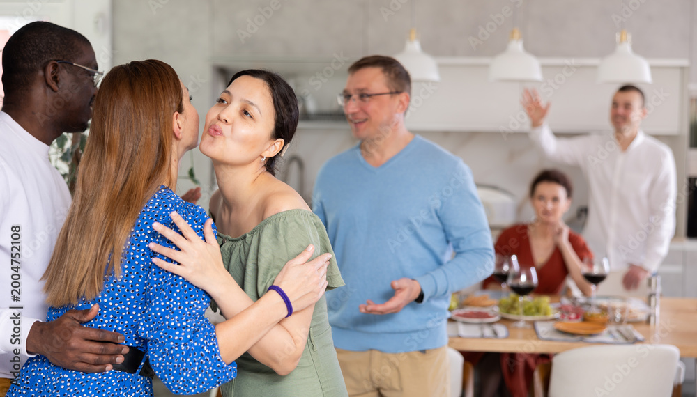 Obraz premium Smiling Asian woman hosting guests at home, greetingly hugging and kissing female friend, enjoying convivial dinner party atmosphere