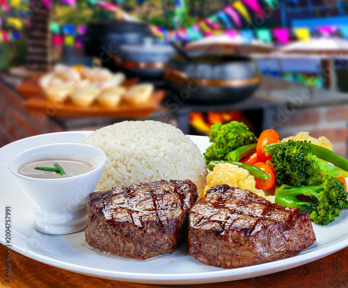 Grilled beef steaks served with white rice, steamed broccoli, carrots, and a side of creamy sauce. Traditional Brazilian outdoor restaurant setting with a wood stove background,