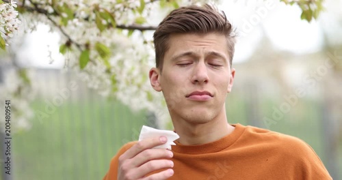 Man with paper tissue suffering from seasonal pollen allergy near blossoming tree on spring day