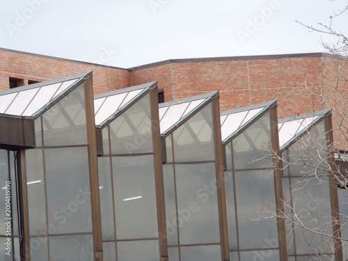 A series of angled, glass sawtooth skylights extend from a modern building with a red brick wall in the background.