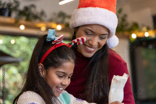 Mother and daughter decorating cookies in kitchen, child holding frosting bag, wearing Santa hat