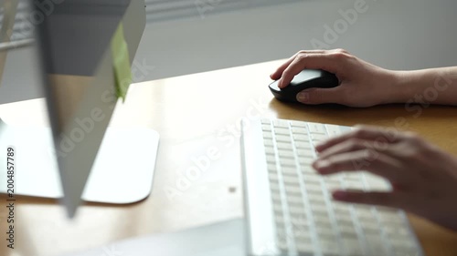 Professional woman using a computer for online work, typing on the keyboard and clicking the mouse on a desk with a monitor and a post-it in an office setting