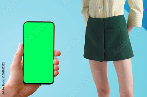  Modern fashion and technology concept featuring a stylish female model in contemporary outfit with striped blouse and green skirt, paired with a hand holding smartphone mockup with blank chroma 