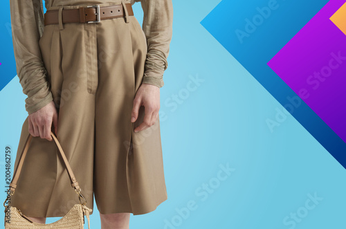  Stylish close-up of a modern beige outfit featuring high-waisted shorts, a tucked-in long sleeve top, and a brown leather belt. The model holds a woven handbag, set against a vibrant geometric blue